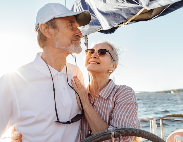 Rijpe vrouw met een zonnebril die haar man omhelst en naar hem kijkt terwijl hij de zeilboot bestuurt