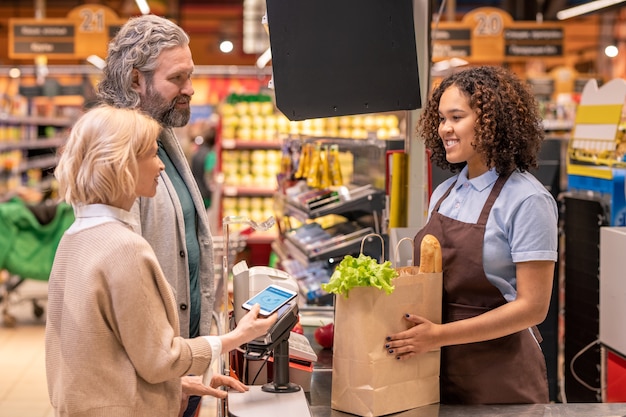 Rijpe vrouw met smartphone betalen voor voedingsproducten in de supermarkt terwijl ze door haar man voor gelukkige jonge kassier staat