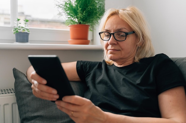 Rijpe vrouw van middelbare leeftijd die thuis op de bank e-boeken behandelt