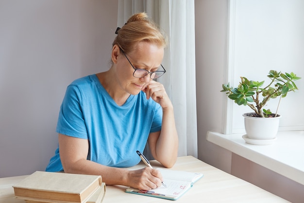 Rijpe vrouw zit met een pen in een dagboek te schrijven terwijl ze thuis aan tafel zit