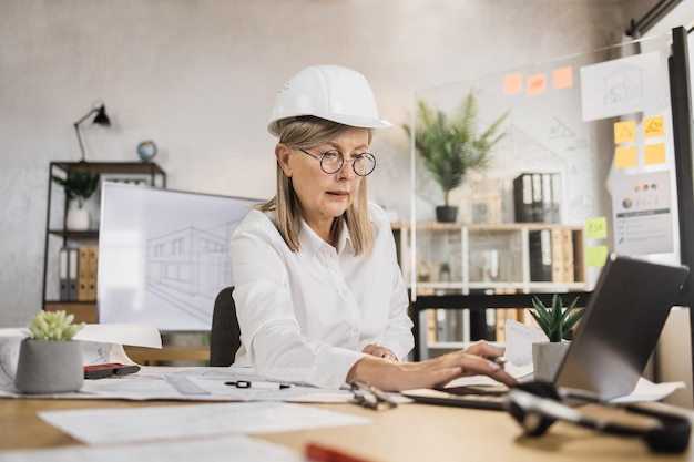 Rijpe vrouwelijke architect die laptopcomputer gebruikt die aan bouwplanschetsen werkt