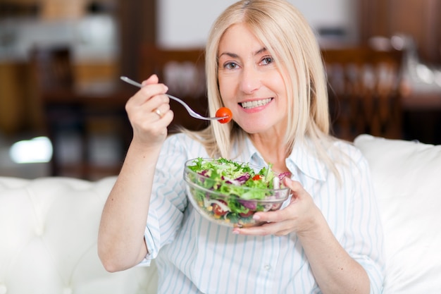 Rijpe vrouwenzitting op een laag thuis terwijl het eten van een salade