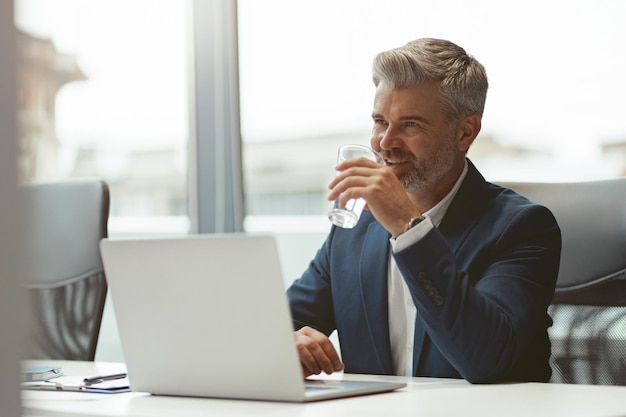 Rijpe zakenman die op laptop werkt en water drinkt terwijl hij in moderne coworking zit