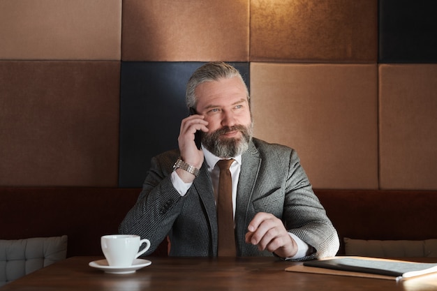 Rijpe zakenman die op mobiele telefoon praat terwijl hij koffie drinkt aan tafel in de coffeeshop