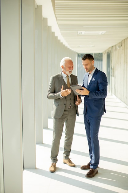 Rijpe zakenman en zijn jonge collega bespreken financieel rapport over digitale tablet in de kantoorgang