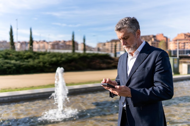 Rijpe zakenman met behulp van smartphone in de buurt van fontein