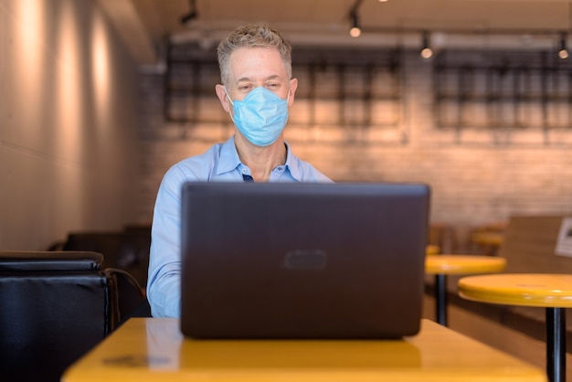 Rijpe zakenman met masker die laptop met behulp van bij de koffiewinkel