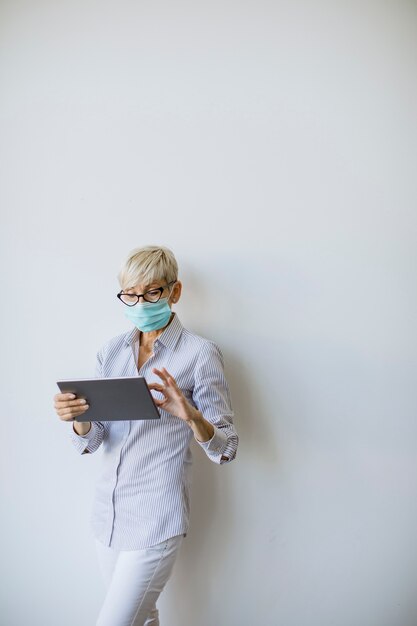 Rijpe zakenvrouw gebruikt een handheld digitale tablet en draagt een masker om infectie door het coronavirus te voorkomen