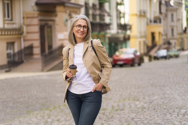 Rijpe zakenvrouw in casual formele greep wegwerpbeker met warme koffie staande buiten de oude stad Rijpe vrouw brengt vrije tijd door met reizen in de Europese stad, genietend van een kopje thee koffie