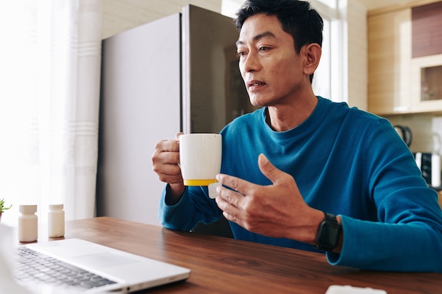 Rijpe zieke man met gelezen ogen hete thee drinken tijdens een videogesprek met arts of collega