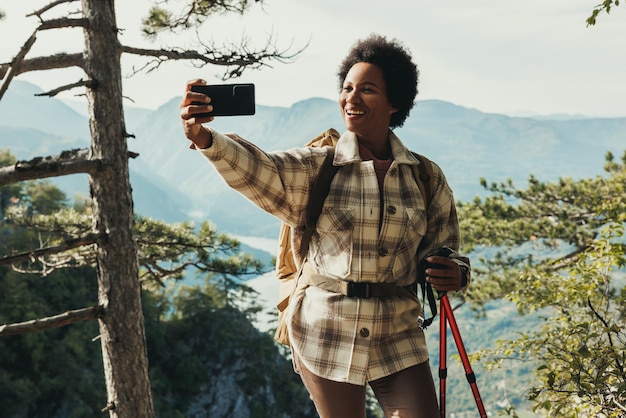 Rijpe zwarte vrouw die een videogesprek voert terwijl ze op de top van een heuvel staat en geniet van het uitzicht tijdens haar wandeling in de bergen.