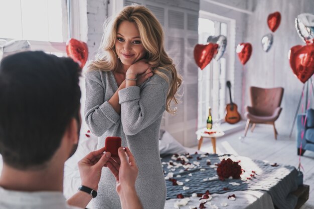 Ring is perfect! Attractive young woman looking at engagement ring with smile while her boyfriend proposing her in the bedroom