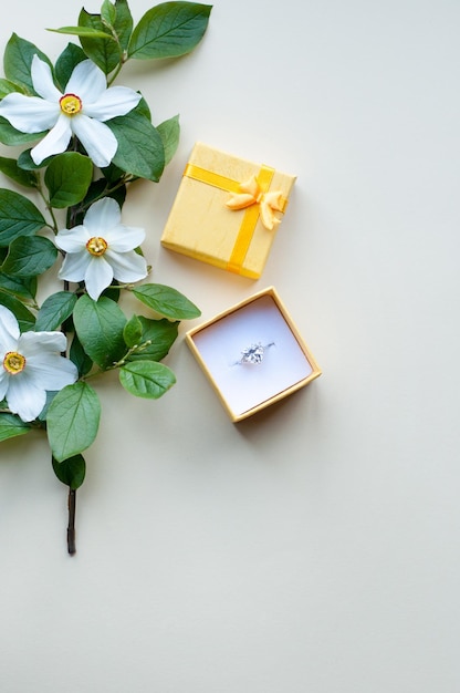 A ring with a large stone in a yellow box next to a branch with narcissus flowers on a beige background