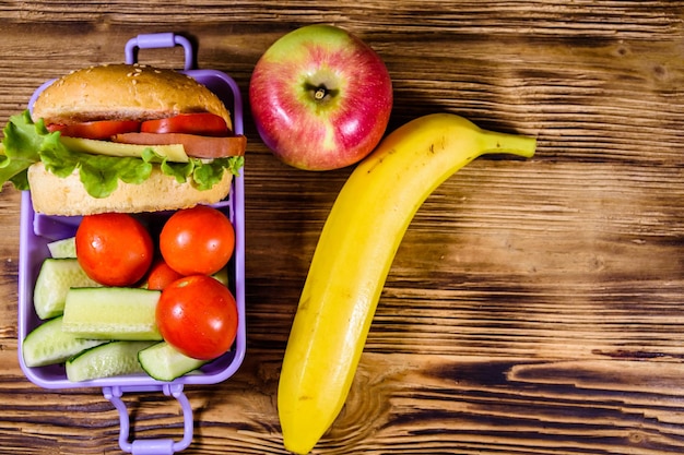 Ripe apple banana and lunch box with hamburger cucumbers and tomatoes on rustic wooden table Top view