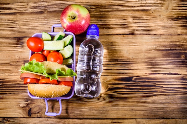 Ripe apple bottle of water and lunch box with hamburger cucumbers and tomatoes on wooden table Top view