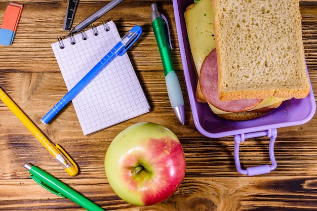 Ripe apple different stationeries and lunch box with sandwiches on a wooden table Top view