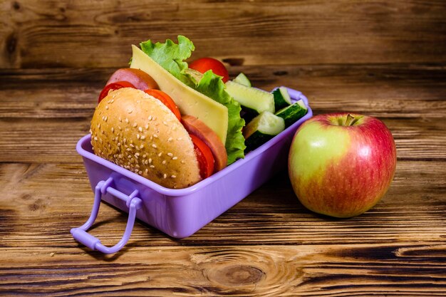 Ripe apple and lunch box with hamburger cucumbers and tomatoes on wooden table