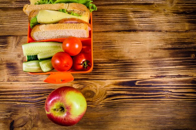 Ripe apple and lunch box with sandwiches cucumbers and tomatoes on rustic wooden table Top view