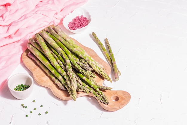 Ripe asparagus on a wooden chopping board. Fresh ingredients ready for cooking healthy food. Modern hard light, dark shadow. White putty background, copy space