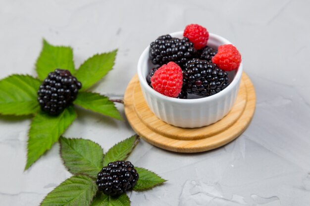 Ripe blackberry with leaves on a wooden cutting board in a white ceramic bowl on concrete background