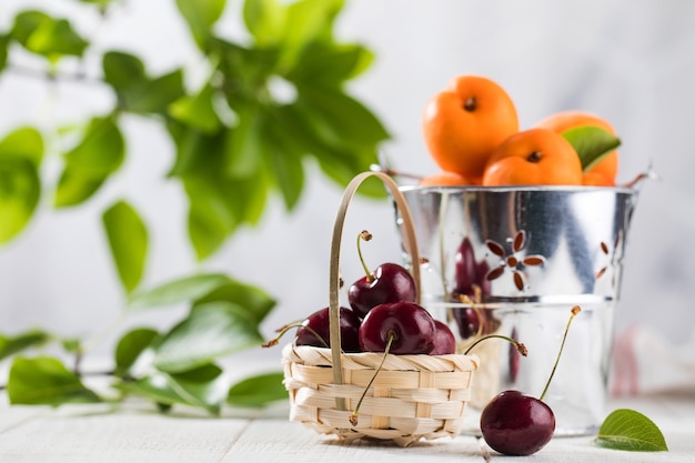 Ripe cherries in a basket and fresh apricots in a bucket on white wooden table natural fresh products
