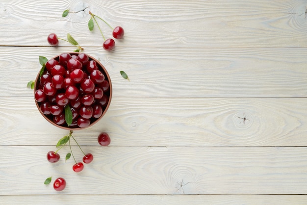 Ripe cherries and leaves in a bowl on a textured wooden background, view from above
