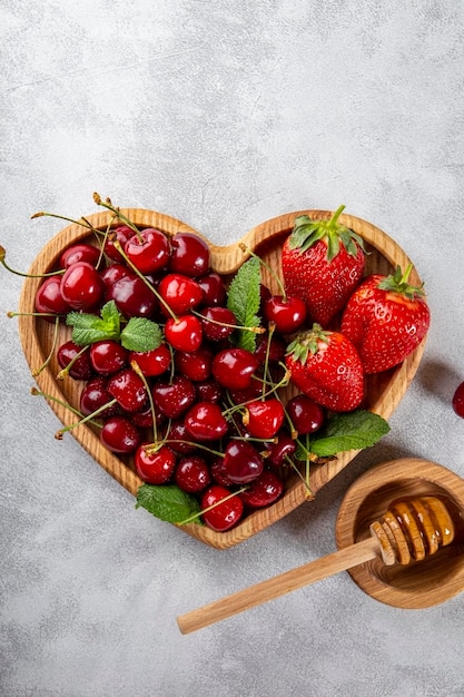 Ripe cherries and strawberries on a wooden plate in the shape of a heart and honey on a light gray