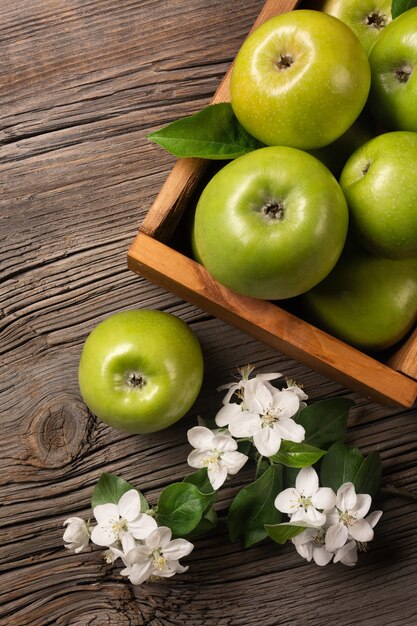 Ripe green apples in wooden box with branch of white flowers on a wooden table. Top view.