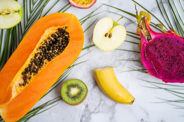 Ripe juicy seasonal tropical fruits. papaya, kiwi, banana,apple, dragon fruit, palm leaves on marble white background. vacation, healthy food, summer concept. top view.