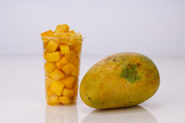 Ripe Mango cubes or cut piece arranged in a transparent glass with mango placed near by on  white color background.