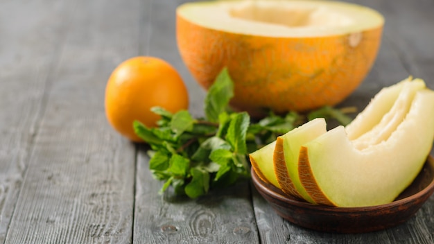 Ripe melon pieces on a clay bowl, banana, mint and orange on a rustic wooden table.