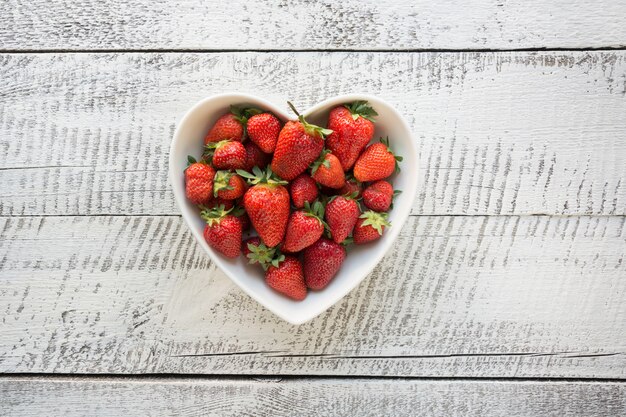 Ripe organic strawberry in plate as heart on white wooden background. Top view.