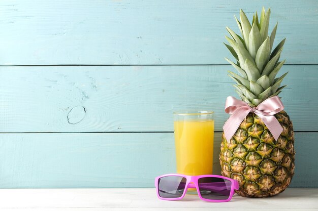 Ripe pineapple fruit with a pink bow, sunglasses and pineapple juice on a blue wooden table. place for text.