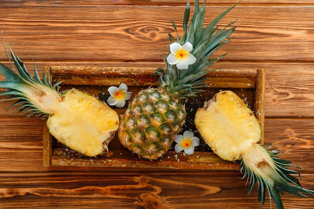 Ripe Pineapple. Tropical summer fruit pineapple halves and whole pineapple on brown dark table in wooden box with tropical plumeria flowers. Flat lay. High quality stock photo