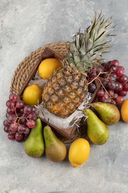 Ripe pineapple in wooden box with various fresh fruits on marble surface. 