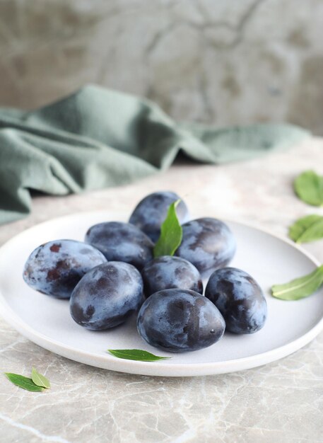 A ripe plum lies in a plate on a marble table Green leaves are scattered around Behind lies a kitchen towel