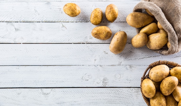 Ripe potatoes in burlap sack and wooden basket freely lying on wooden board.