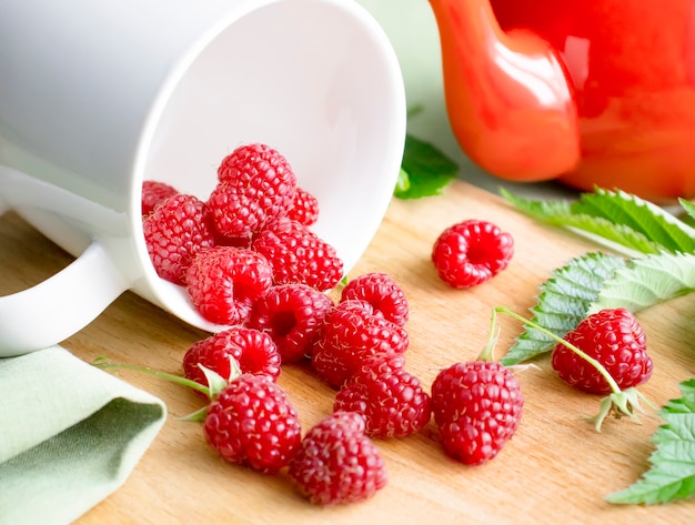 Ripe raspberry berries on a wooden background are poured out of a white cup.