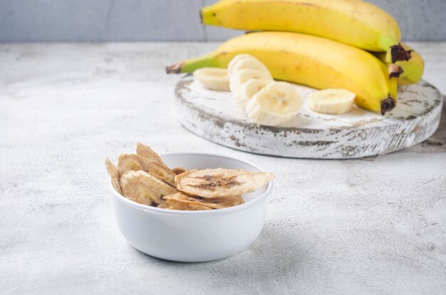 Спелые сырые бананы и сушеные банановые ломтики чипсов в тарелке на светло-сером фоне. Фруктовые чипсы. Концепция здорового питания, закуски, без сахара. Вид сверху, копия пространства.