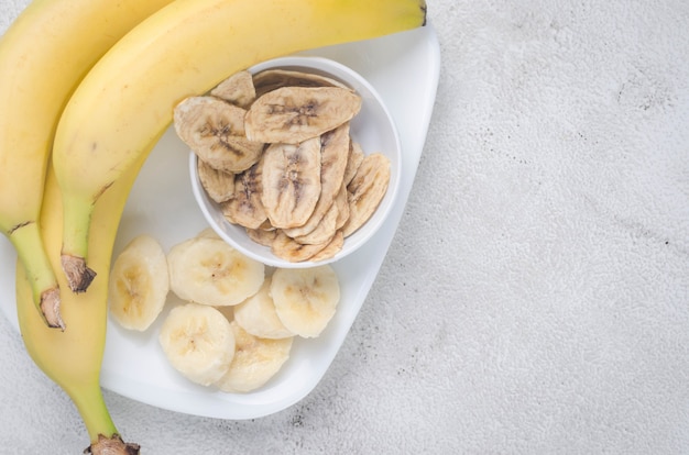 La banana cruda matura e le fette di banana essiccate scheggiano nel piatto su fondo grigio chiaro chips di frutta.