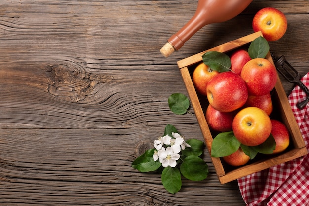 Ripe red apples in wooden box with branch of white flowers, glass and bottle of cider on a wooden table. Top view with space for your text.