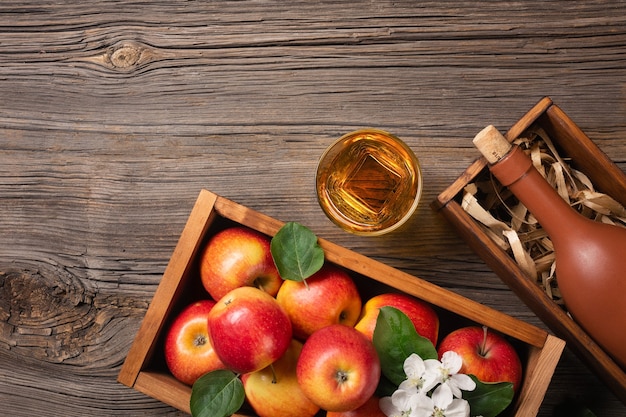 Ripe red apples in wooden box with branch of white flowers,\
glass and bottle of cider on a wooden table. top view with space\
for your text.