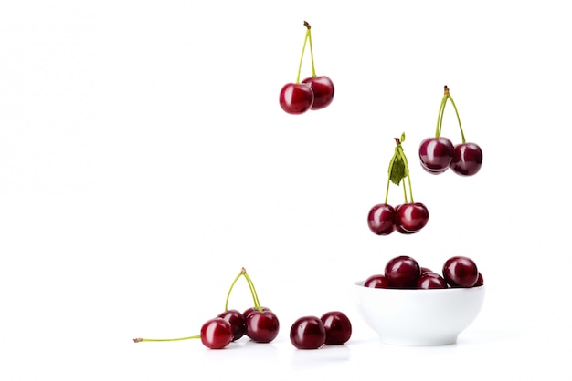 Ripe red cherries with green sticks and leaves fall into a white ceramic bowl.