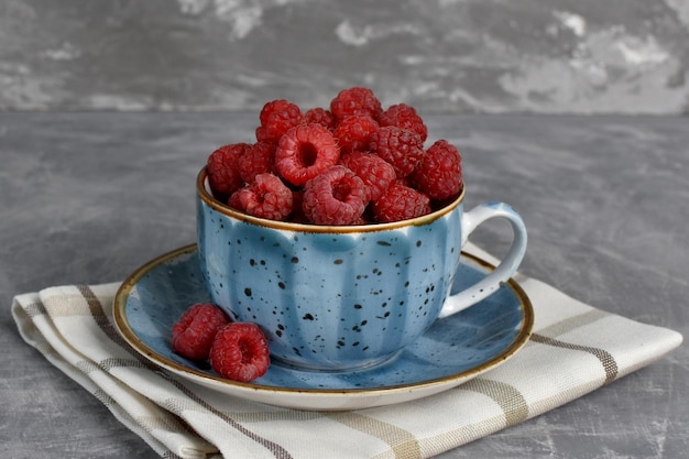 Photo ripe red raspberries in a beautiful blue cup on a gray stone background summer berries