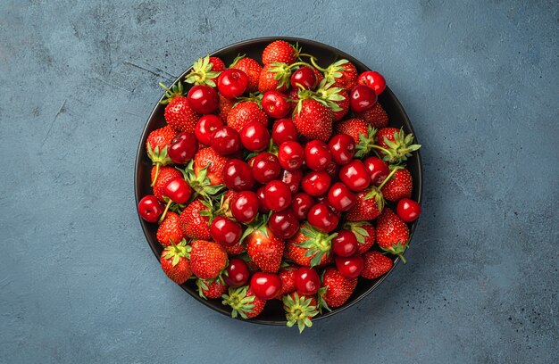 Ripe strawberries and cherries in a black plate on a dark background. Top view, copy space.