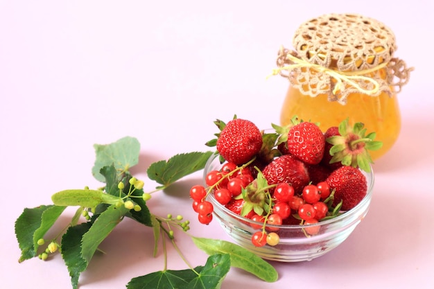 Ripe strawberries and red currants in a glass cup a jar of jam a linden branch on a white background side view a place for text