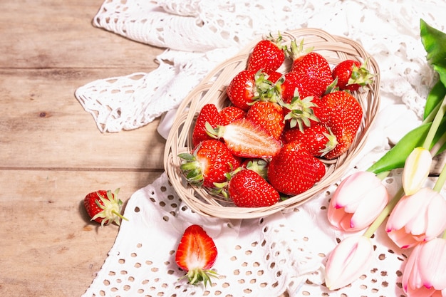 Ripe strawberries in wicker plate. Fresh berries, aromatic mint, pink tulips. Old wooden table, vintage crocheted napkin, rustic style, copy space