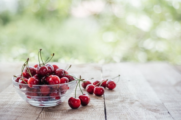 Ripe sweet cherries in a glass bowl on wooden table with green bokeh background, summer fruits