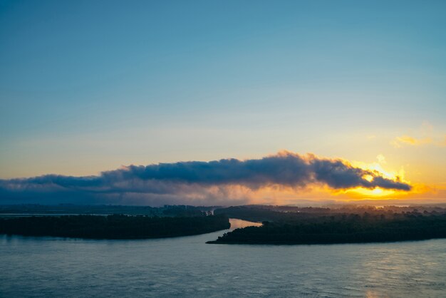 Riverbank with forest and fog. Orange glow from picturesque dawn reflected in river. 