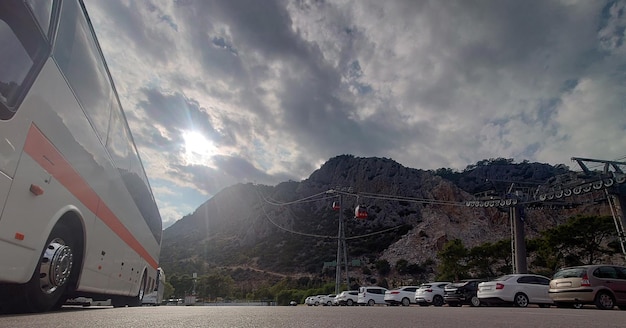 Road ground level view of a parked luxury tourist coach in a parking lot with cars below a mountain peak with cableway to the top for sightseeing
